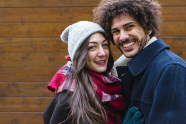Porträt eines glücklichen jungen Paares in Winterkleidung vor einer Holzwand - MGIF00315