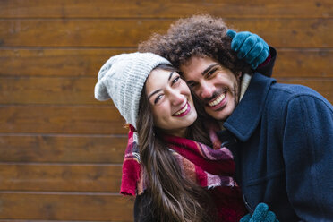 Portrait of happy young couple in winterwear hugging in front of wooden wall - MGIF00316