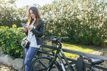 Lächelnde junge Frau mit Fahrrad im Park, die ein Mobiltelefon benutzt - KIJF02362