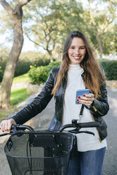 Porträt einer lächelnden jungen Frau mit Fahrrad und Mobiltelefon im Park - KIJF02365