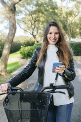 Portrait of smiling young woman with bicycle and cell phone in park - KIJF02365