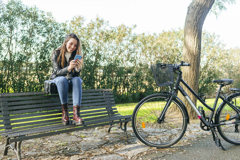 Junge Frau mit Fahrrad, die auf einer Bank im Park sitzt und ein Mobiltelefon benutzt - KIJF02367