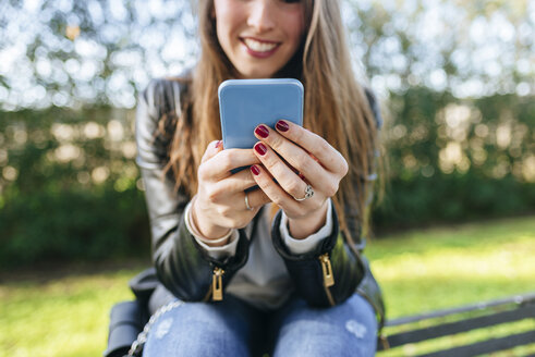 Nahaufnahme einer jungen Frau, die auf einer Bank im Park sitzt und ein Handy benutzt - KIJF02368