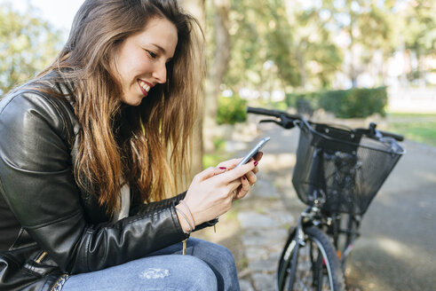 Junge Frau mit Fahrrad, die im Park sitzt und ein Mobiltelefon benutzt - KIJF02369