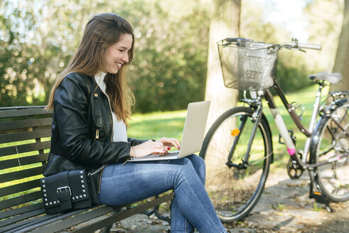 Junge Frau sitzt auf einer Bank im Park und benutzt einen Laptop - KIJF02370