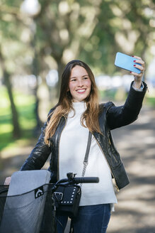 Lächelnde junge Frau mit Fahrrad im Park macht ein Selfie - KIJF02374