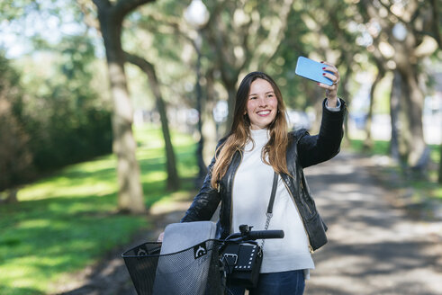 Lächelnde junge Frau mit Fahrrad im Park macht ein Selfie - KIJF02375