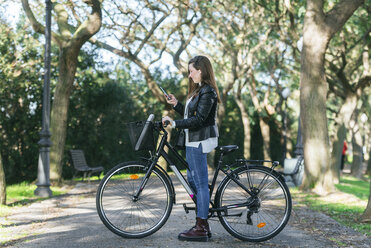 Junge Frau mit Fahrrad im Park, die ein Mobiltelefon benutzt - KIJF02379