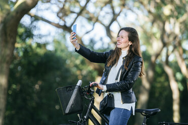 Lächelnde junge Frau mit Fahrrad im Park macht ein Selfie - KIJF02380