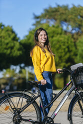 Portrait of smiling young woman with bicycle and headphones in park - KIJF02384