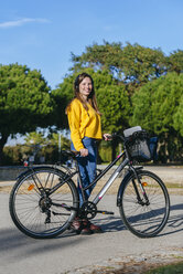Porträt einer lächelnden jungen Frau mit Fahrrad und Kopfhörern im Park - KIJF02385