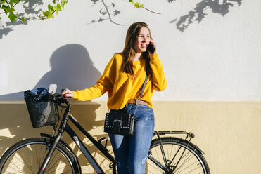 Young woman with bicycle talking on cell phone at a wall - KIJF02391
