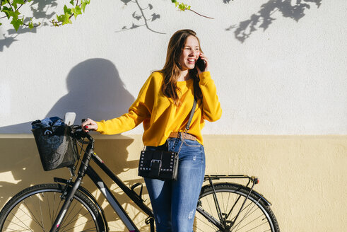 Junge Frau mit Fahrrad im Gespräch mit Handy an einer Wand - KIJF02391