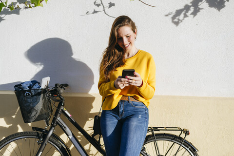 Lächelnde junge Frau mit Fahrrad und Mobiltelefon an einer Wand, lizenzfreies Stockfoto