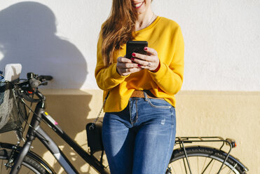 Close-up of young woman with bicycle using cell phone at a wall - KIJF02393