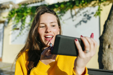 Junge Frau trägt Lippenstift auf und betrachtet sich selbst auf dem Handy - KIJF02394