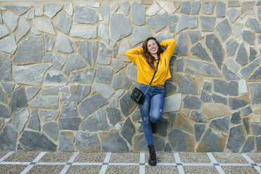 Porträt einer glücklichen jungen Frau, die sich gegen eine Steinmauer lehnt - KIJF02396