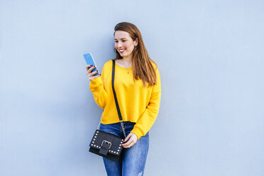 Lächelnde junge Frau, die an einer Wand steht und ein Mobiltelefon benutzt - KIJF02401