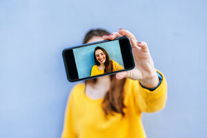 Lächelnde junge Frau macht ein Selfie vor einer blauen Wand - KIJF02403