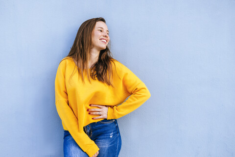 Glückliche junge Frau mit gelbem Pullover an blauer Wand stehend, lizenzfreies Stockfoto