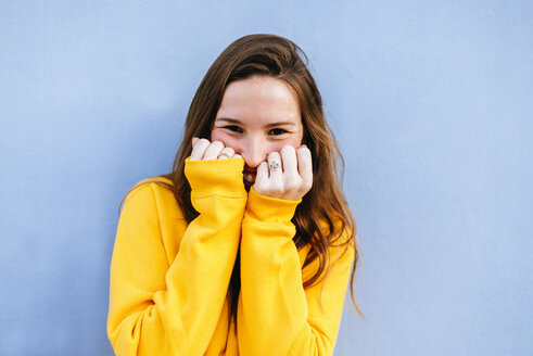 Porträt einer glücklichen jungen Frau mit gelbem Pullover an einer blauen Wand - KIJF02405