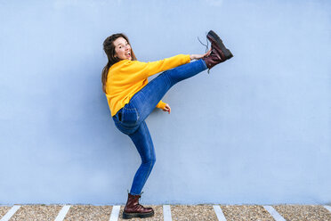Verspielte junge Frau mit erhobenem Bein an blauer Wand - KIJF02406