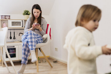 Frau sitzt in der Küche und benutzt ihr Smartphone, während ihre kleine Tochter im Vordergrund steht - KMKF00770