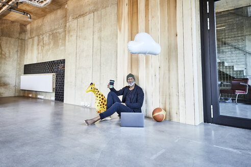Entspannter Geschäftsmann auf dem Boden sitzend mit Kaffee, Laptop, Ball und Giraffenfigur - FMKF05424