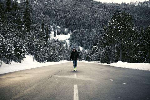 Junger Mann geht auf einer verschneiten Straße mit Bäumen im Hintergrund, lizenzfreies Stockfoto