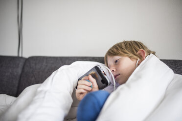 Junge mit Decke, der ein Mobiltelefon benutzt, während er zu Hause auf dem Sofa an der Wand liegt - CAVF62326