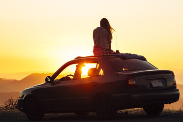 Rückansicht einer Frau, die auf dem Autodach sitzend die Aussicht gegen den klaren Himmel bei Sonnenuntergang betrachtet - CAVF62360