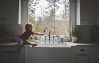 Seitenansicht eines niedlichen Mädchens, das seine Hand im Waschbecken wäscht, während es auf der Küchentheke vor dem Fenster sitzt - CAVF62386