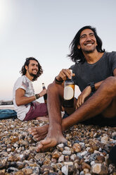 Lächelnde Freunde mit Alkoholflaschen sitzen auf Kieselsteinen am Strand gegen den Himmel - CAVF62401