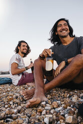 Smiling friends with alcohol bottles sitting on pebbles at beach against sky - CAVF62401