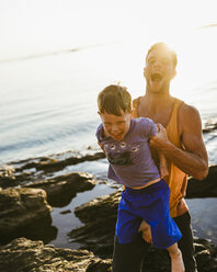 Happy father carrying son while standing on rocks at beach against sky during sunset - CAVF62427