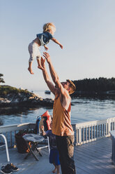 Glücklicher Vater wirft seinen Sohn, während er auf dem Steg über dem See steht, gegen den klaren Himmel während des Sonnenuntergangs - CAVF62429