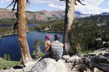 Rear view of couple looking at lake while sitting on rocks during sunny day - CAVF62439