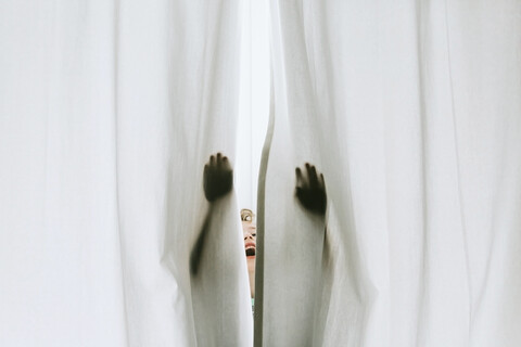 Fröhlicher Junge spielt hinter weißem Vorhang zu Hause, lizenzfreies Stockfoto