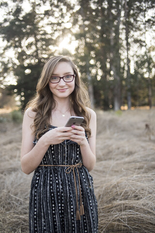 Porträt einer lächelnden jungen Frau, die ein Smartphone benutzt, während sie gegen Bäume im Wald steht, lizenzfreies Stockfoto