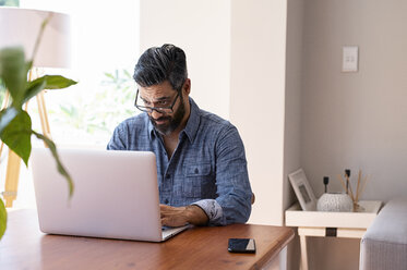 Geschäftsmann mit Brille, der einen Laptop-Computer auf einem Holztisch benutzt, während er zu Hause sitzt - CAVF62489