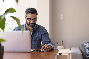 Businessman with laptop computer on wooden table using mobile phone while sitting at home - CAVF62491