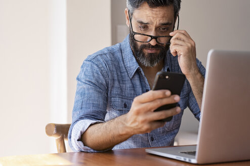 Geschäftsmann mit Laptop auf einem Holztisch, der ein Smartphone benutzt, während er zu Hause an der Wand sitzt - CAVF62493