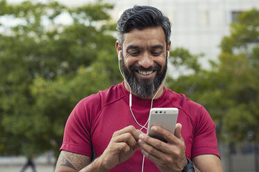 Lächelnder, selbstbewusster Mann, der sein Smartphone benutzt, während er im Park vor Bäumen steht - CAVF62502