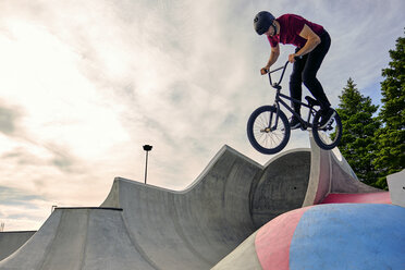 Male rider with BMX bike jumping on concrete ramp against cloudy sky at skateboard park - CAVF62564