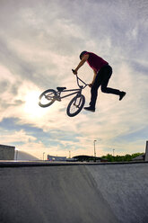 Low angle view of male rider with BMX bike performing tailwhip on concrete ramp against cloudy sky - CAVF62566