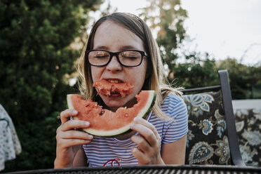 Mädchen mit Brille isst Wassermelone, während sie auf einem Stuhl gegen Bäume im Garten sitzt - CAVF62604
