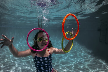 Smiling girl wearing swimming goggles while playing with colorful rings in pool - CAVF62608