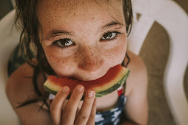 Hochwinkliges Porträt eines nassen Mädchens, das auf einem Stuhl sitzend eine Wassermelone isst - CAVF62610