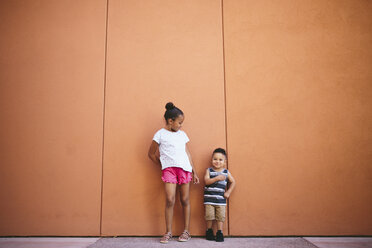 Geschwister in voller Länge auf dem Fußweg vor der orangefarbenen Wand stehend - CAVF62615
