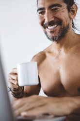 Niedriger Blickwinkel auf einen lächelnden Mann ohne Hemd, der eine Kaffeetasse hält, während er einen Laptop-Computer zu Hause benutzt - CAVF62630